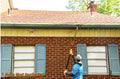 A person using an Extend-A-Reach telescoping pole with utility hook to hang string lights along a house roofline. This Christmas light pole allows users to decorate safely from the ground without a ladder. The image includes the text “Pole Not Included.