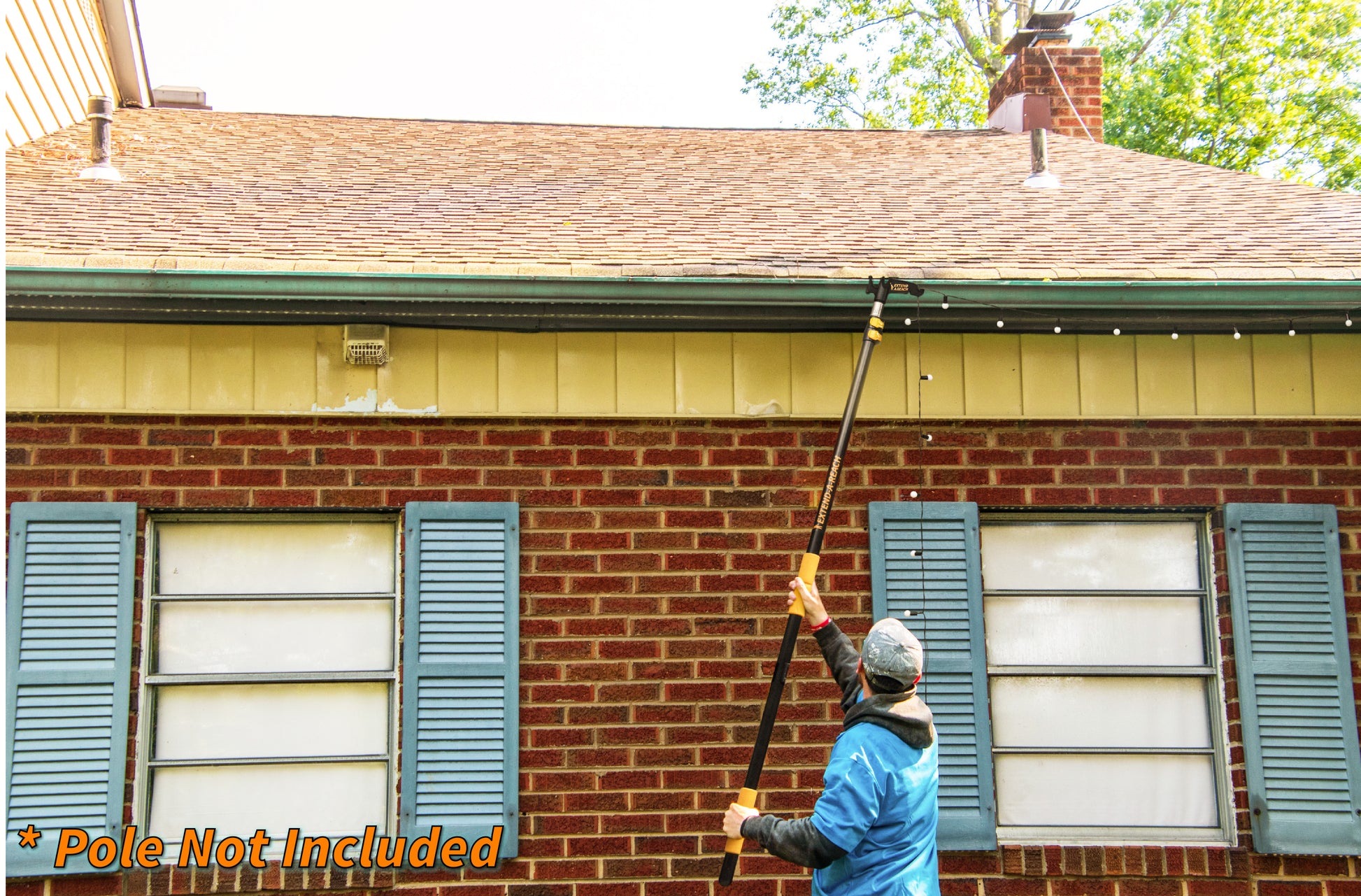 A person using an Extend-A-Reach telescoping pole with utility hook to hang string lights along a house roofline. This Christmas light pole allows users to decorate safely from the ground without a ladder. The image includes the text “Pole Not Included.