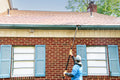 A person using an Extend-A-Reach telescoping pole with utility hook to hang string lights along a house roofline. This Christmas light pole allows users to decorate safely from the ground without a ladder.