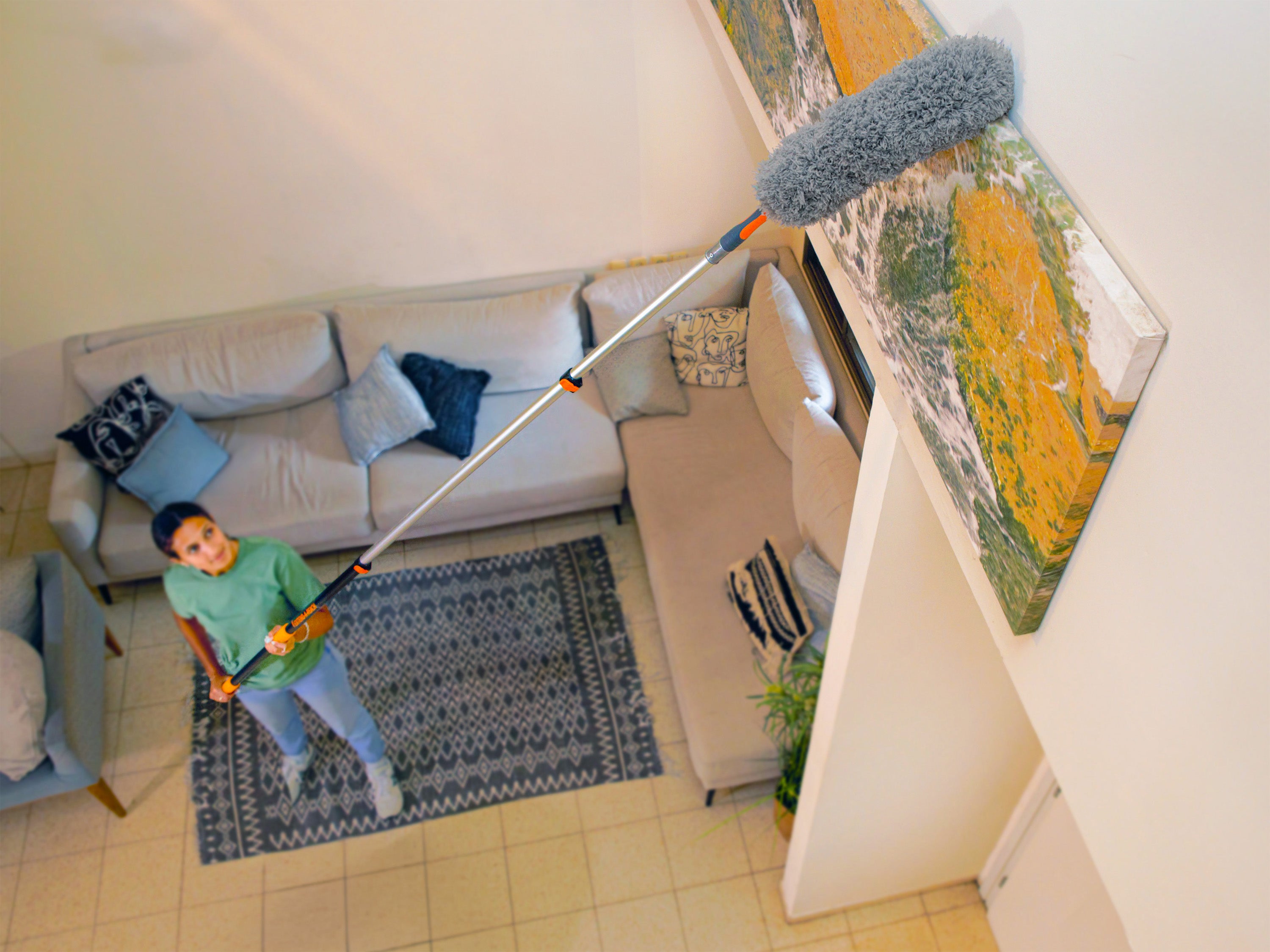 Woman using the Extend-A-Reach telescoping extension pole with feather duster attachment to clean a high wall-mounted painting in her living room - a long reach duster designed for high ceilings and hard-to-reach indoor areas.